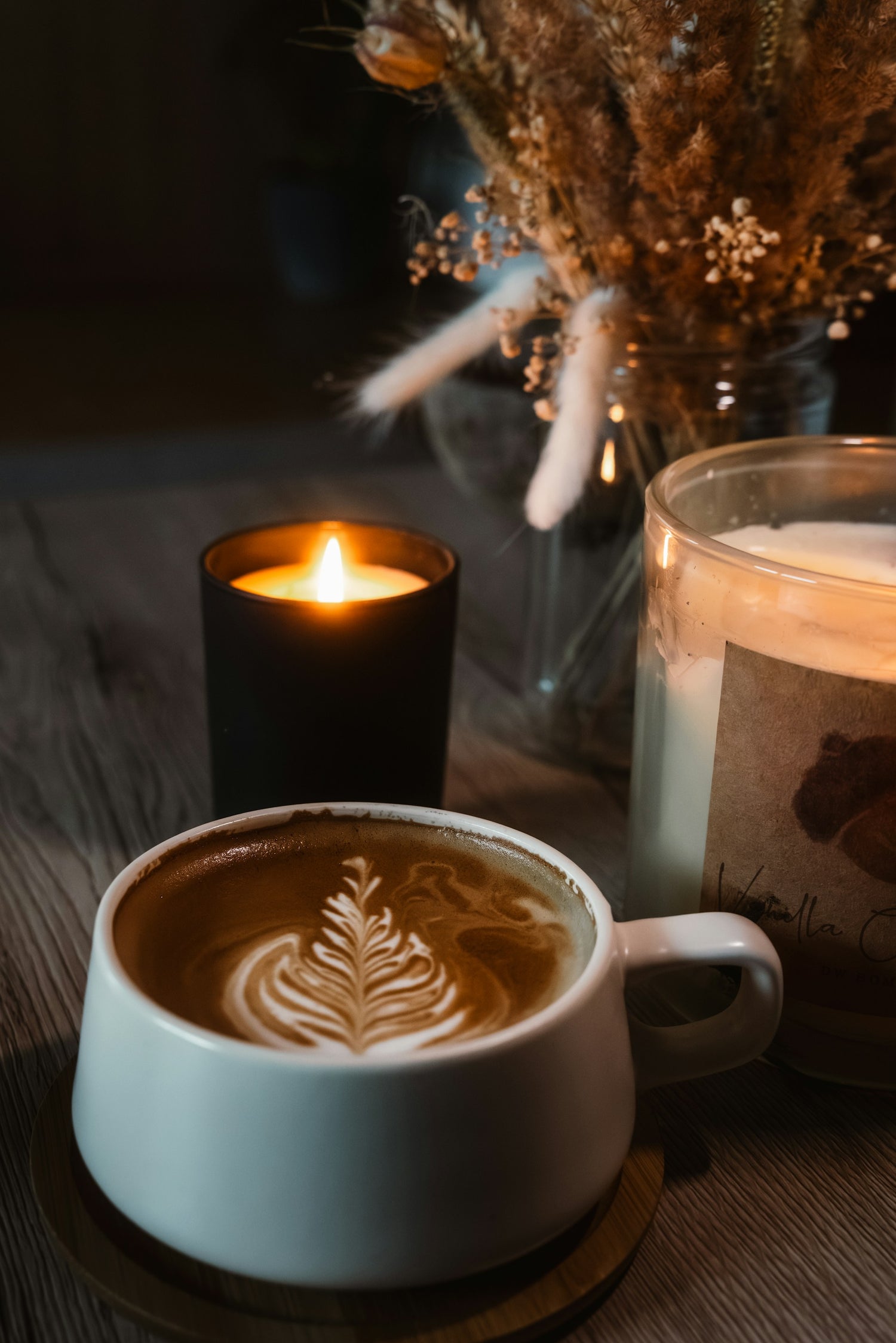 Cup of coffee with latte art, lit candle, and dried flowers on a dark surface